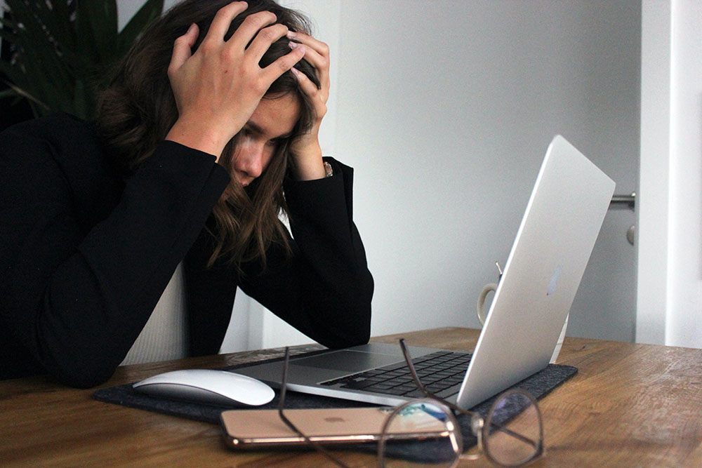 A woman with her hands on her head, glasses placed to the side, staring in frustration at a MacBook